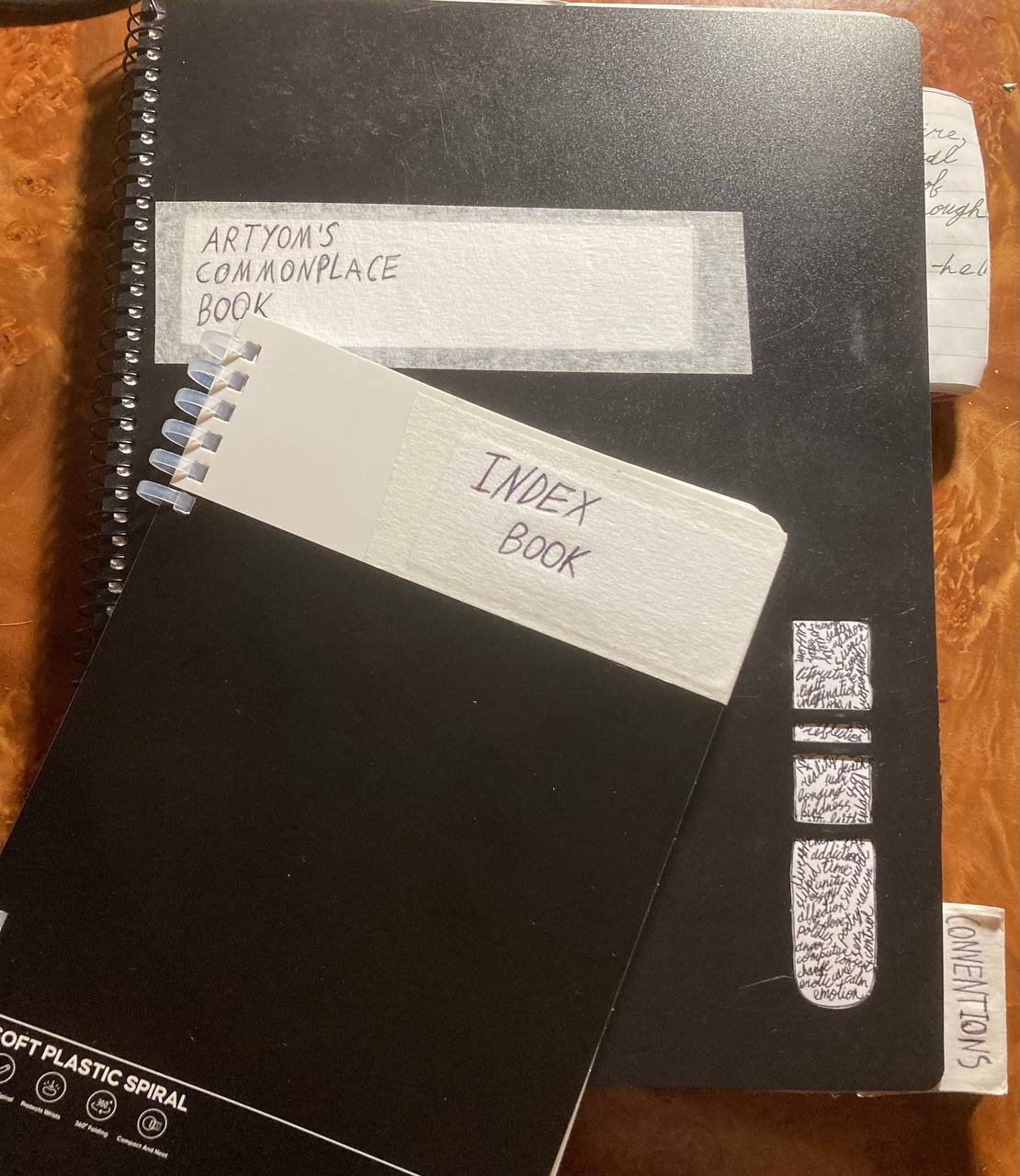 Two notebooks placed atop each other, so that labels on both can be seen. On these, the black ink says “Artyom’s Commonplace Book” (on a larger one,) and “Index Book” (on a smaller one.) Commonplace Book is a spiral-bound plastic cover A4 notebook that has somewhat thin paper, with glued-in pieces of older notebooks sometimes protruding on the side of it. The cover has a nice uneven texture of an unpolished plastic. The Index Book has a silicone spiral bind and cardboard cover. Both labels are made with paper tape that has a slightly rugged texture and nice creamy color.