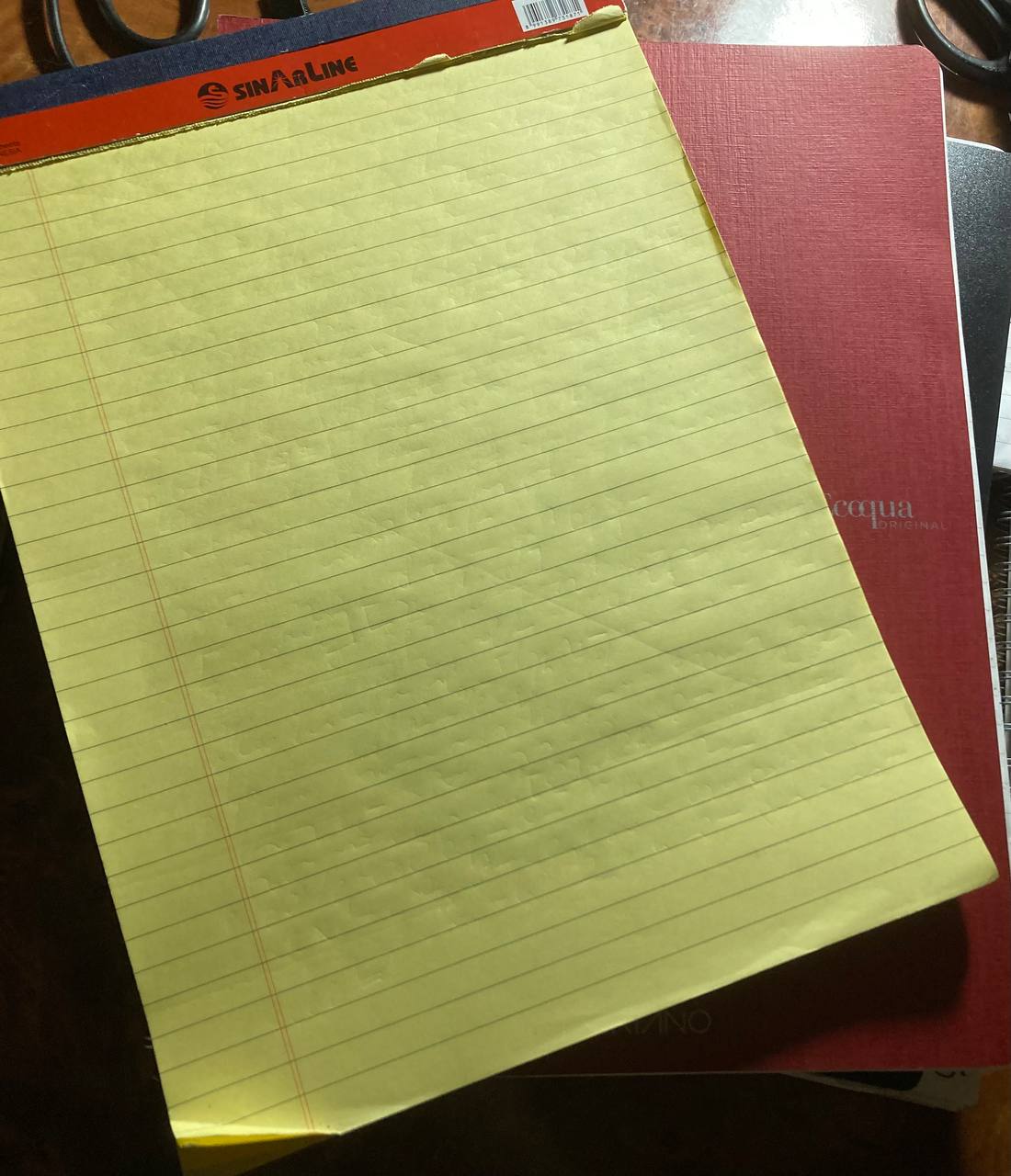 Photo of two notebooks. The one below is red and fancy, spiral-bound, with good paper. The one in the front has no cover, yellow pages, and is a vertical tear-off design.