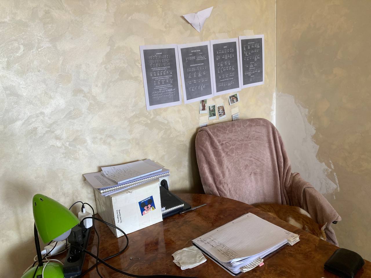 Photo of a dining table in a moderately cringe Armenian apartment. Behind it, there is a chair with a pink blanket over it (yes, even in summer!) On the table, there’s an open notebook and some wires. In the corner of the table, there’s a wine box set on the side, used as some kind of a shelf, with more notebooks and laptops protruding from it.